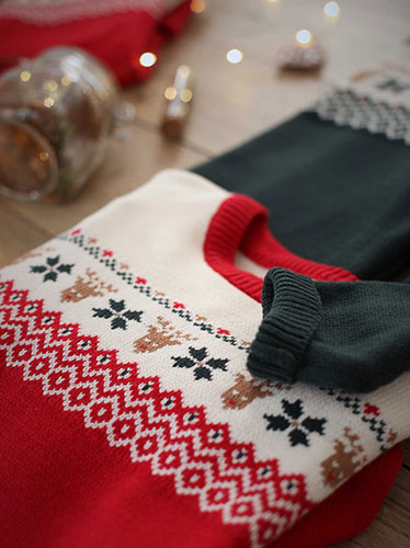 Photographie en gros plan d'un pull de Noël rouge et blanc à motifs de rennes et de flocons, avec des vêtements verts pliés et des lumières de Noël en arrière-plan.
