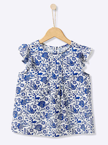 Photographie de produit d'une blouse blanche pour enfant à motifs bleus de fleurs et d'oiseaux, suspendue à un cintre en bois sur fond clair.