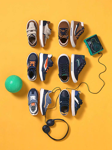 Photographie en vue de dessus de six paires de baskets pour enfant, disposées sur un fond jaune avec un ballon vert et des accessoires audio.