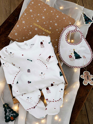 Photographie d'un coffret cadeau Vertbaudet contenant des vêtements blancs à motifs de Noël pour bébé, accompagné d'un bavoir, de décorations et d'une guirlande lumineuse.