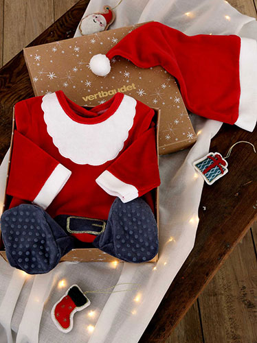 Coffret cadeau de Noël Vertbaudet contenant une tenue de Père Noël pour bébé, avec bonnet rouge, présenté sur une table en bois avec des décorations et une guirlande lumineuse.
