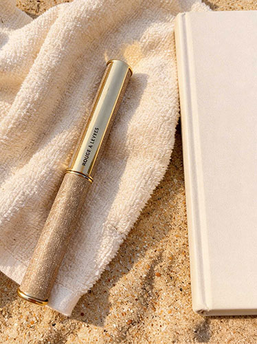 Rouge à lèvres dans un étui doré posé sur une serviette beige, sur le sable, à côté d'un livre blanc, sous une lumière naturelle ensoleillée.
