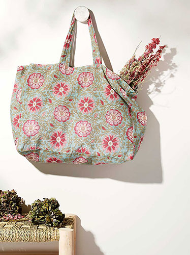 Photographie d'un grand sac cabas en tissu bleu clair à motifs floraux roses, suspendu à un crochet mural, avec un bouquet de fleurs séchées.