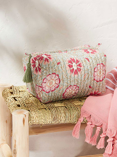 Photographie d'une trousse matelassée à motifs floraux roses sur fond bleu clair, posée sur un banc en fibres tressées à côté d'un tissu rose à franges.