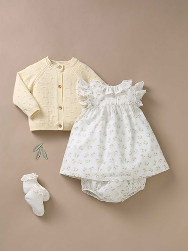 Photographie à plat d'une tenue pour bébé composée d'un gilet en maille écru, d'une robe blanche à motifs fleuris, d'un bloomer assorti et de chaussettes sur fond beige.