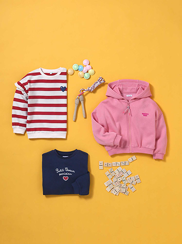 Photographie à plat de vêtements pour enfants, avec un sweat rayé rouge et blanc, un gilet rose et un sweat bleu marine, accompagnés de jouets sur fond jaune.
