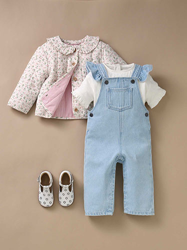 Photographie à plat d'une tenue pour bébé composée d'une salopette en jean bleu clair, d'un tee-shirt blanc, d'une veste fleurie et de petites chaussures blanches sur fond beige.